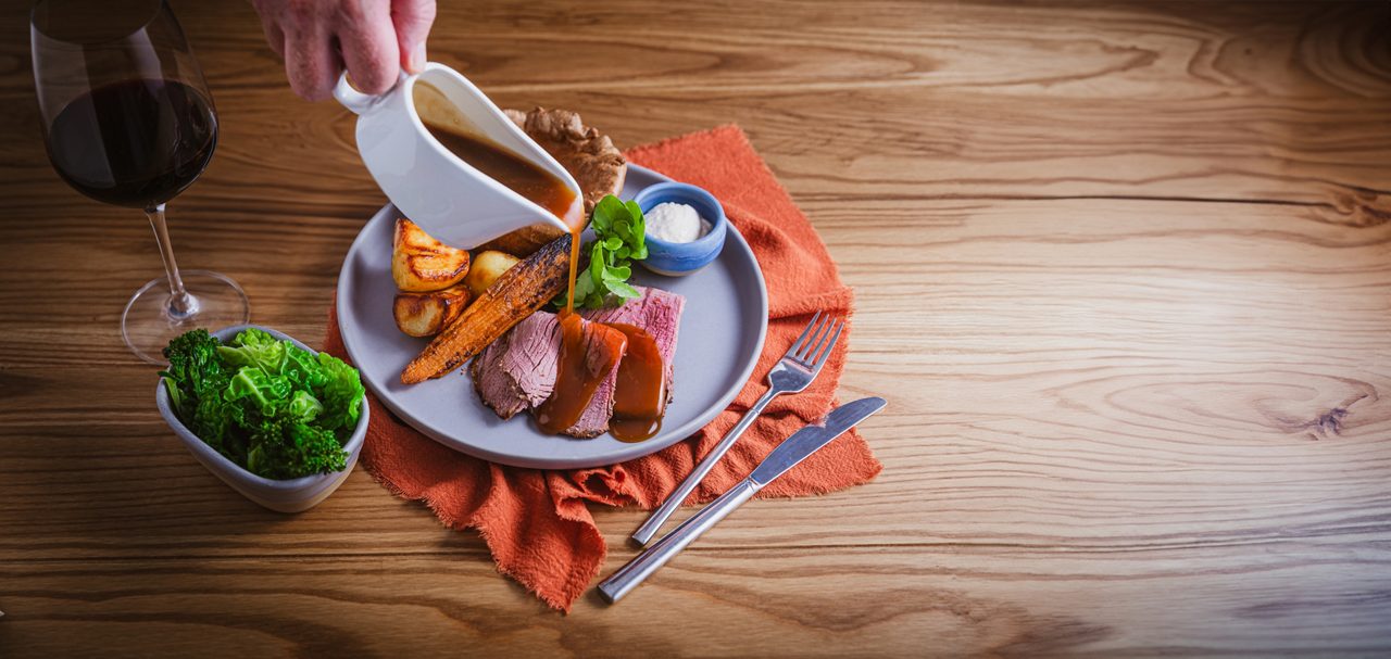 Roast beef slices receive gravy from a white jug, with roast potatoes, carrot, Yorkshire pudding, and horseradish, on a wooden table beside a napkin, greens bowl, and red wine.